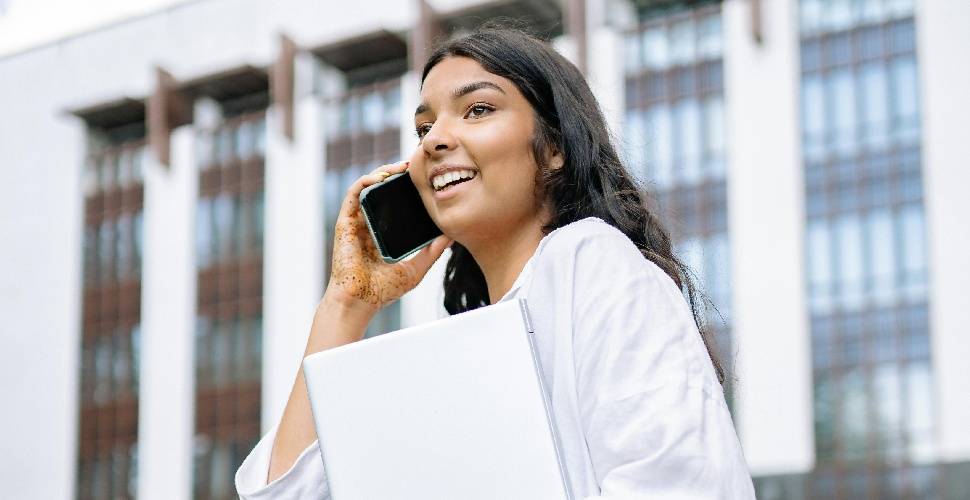 A woman speaking on a mobile phone, holding a file, in an outdoor setting in front of some buildings