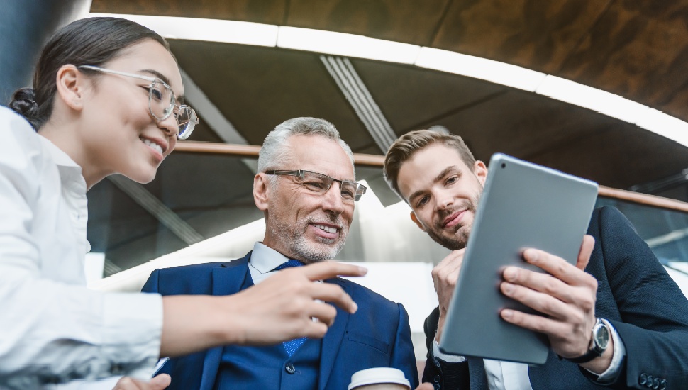 Three professionals of different ages collaborating in an office: a younger woman, a middle-aged man, and an older man with grey hair, all looking at a tablet together. 