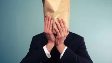 Person in a suit with a paper bag over their head against a blue background.
