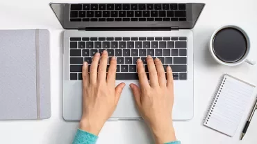 Hands typing on a laptop next to a notebook and coffee cup.