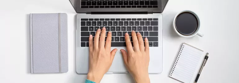 Hands typing on a laptop next to a notebook and coffee cup.