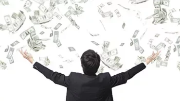 A man in a suit stands with arms raised as dollar bills rain down around him.