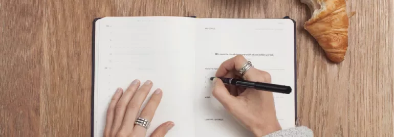 Person writing in a notebook with a pen, croissant on table.