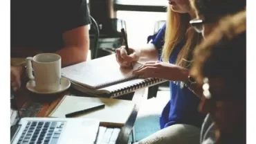 People discussing work around a table with notebooks and a laptop.