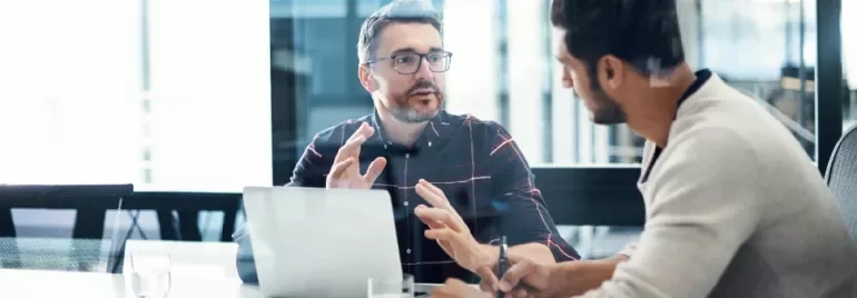 Two men discussing at a table with a laptop in an office setting.