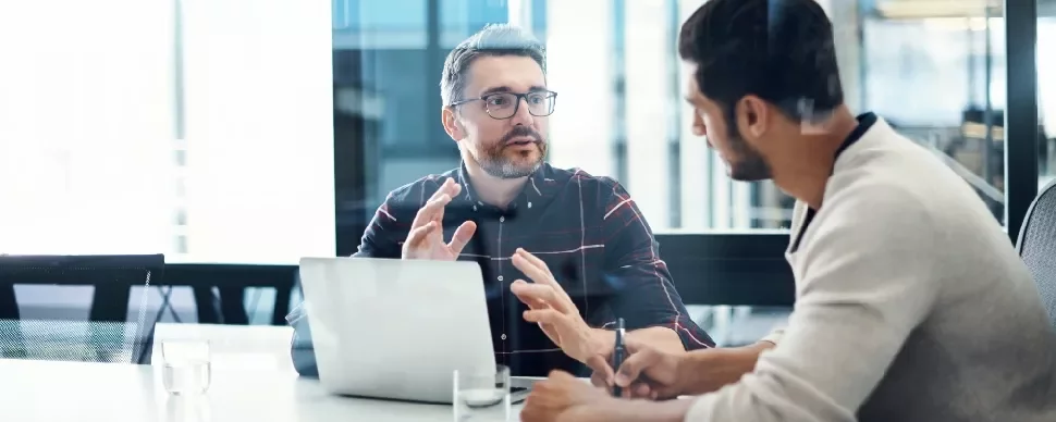 Two men discussing at a table with a laptop in an office setting.