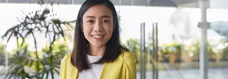 Smiling woman in yellow jacket standing indoors with plants in the background.