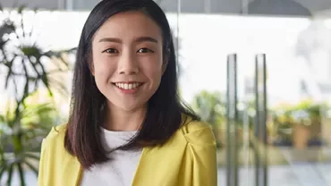 Smiling woman in yellow jacket standing indoors with plants in the background.