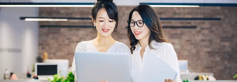 Two women smiling and looking at a laptop in an office environment.