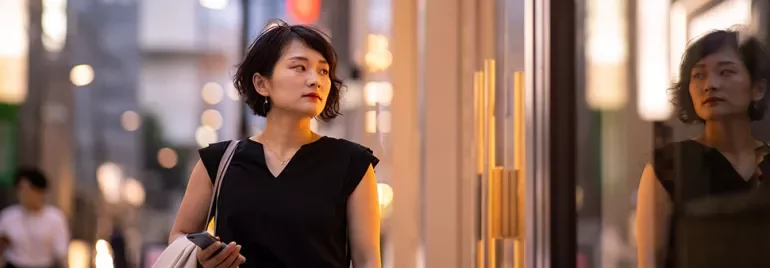 Woman walking past a storefront, holding a bag and phone.