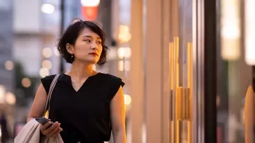 Woman walking past a storefront, holding a bag and phone.