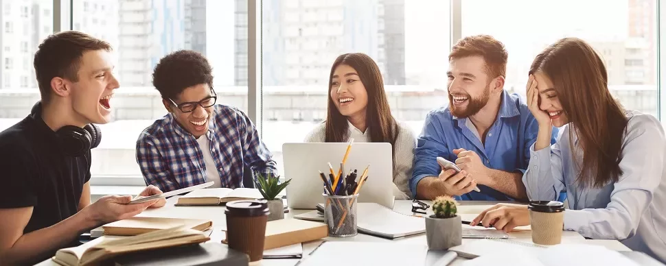 Diverse group of students laughing and studying together with laptops and coffee in a modern setting.