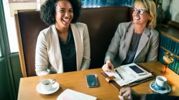 Two women smiling and chatting at a cafe table with papers and coffee.