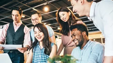 A group of six colleagues smiling around a laptop in an office.
