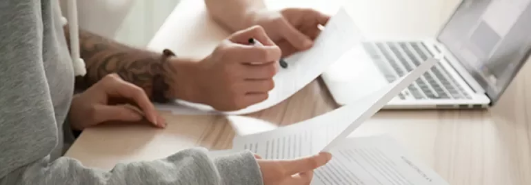 Two people reviewing documents at a desk with a laptop in the background.