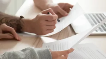 Two people reviewing documents at a desk with a laptop in the background.