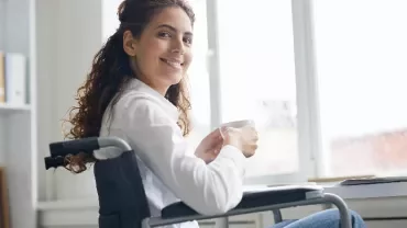 Woman in a wheelchair smiling while holding a mug by a window.