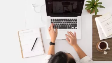 Overhead view of person working on a laptop with a planner, plant, and cup of coffee on the desk.