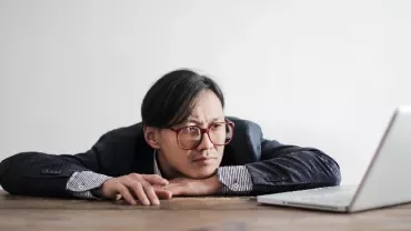 Man in suit and glasses staring thoughtfully at an open laptop on a wooden table.