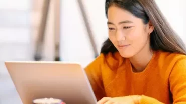 Woman in orange sweater working on a laptop at a desk.