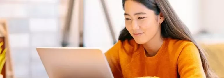 Woman in orange sweater working on a laptop at a desk.