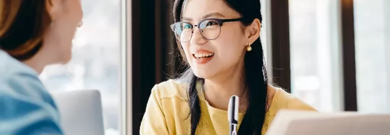 Two women having a conversation at a table, one holding a pen and smiling.