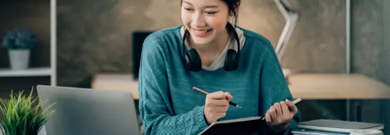 Woman smiling, taking notes at a desk with a laptop and headphones.