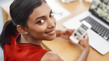 Woman in red top smiling, holding a smartphone while looking at a laptop on a wooden desk.