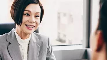 Woman in a gray suit talking to another person in an office setting.