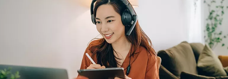 Smiling woman taking notes on a tablet while wearing headphones in a cozy room.