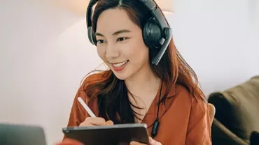 Smiling woman taking notes on a tablet while wearing headphones in a cozy room.