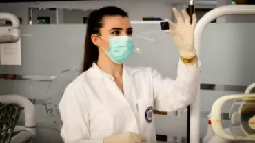 Scientist in lab coat and mask examining a small object in a laboratory.