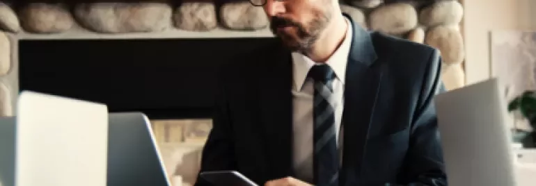 Man in suit working on laptop and smartphone at a table with a coffee cup.