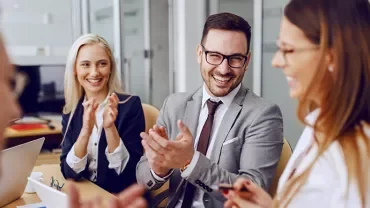 A group of business professionals sitting at a table, laughing and clapping during a meeting.