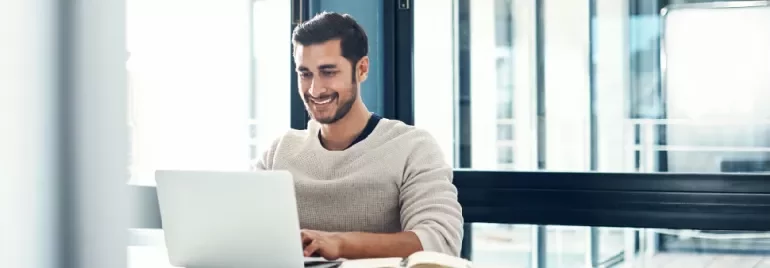 Man smiling while using a laptop at a desk with a notebook open.