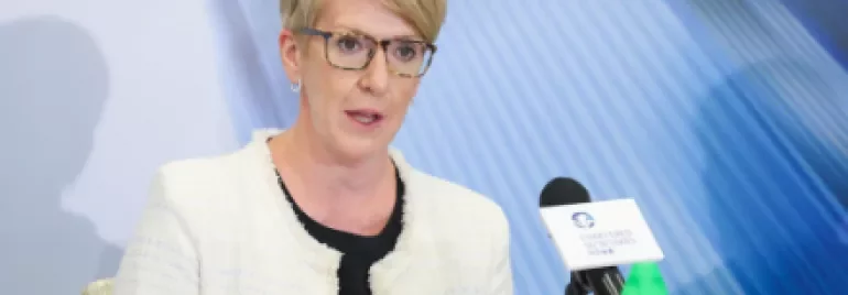 A woman with short blond hair and glasses speaks at a conference, seated with a tablet and microphone on the table.