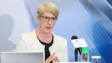 A woman with short blond hair and glasses speaks at a conference, seated with a tablet and microphone on the table.