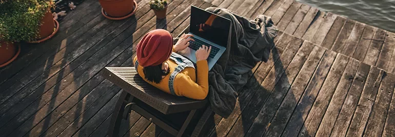 Person in a red hat using a laptop on a wooden deck near water.