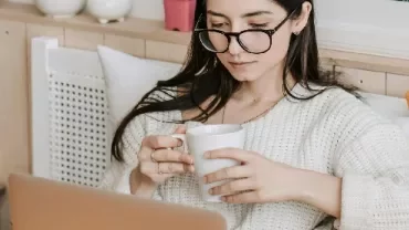 Woman in glasses reading on a laptop and holding a mug
