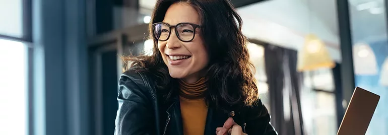Woman in glasses smiling while seated indoors using a laptop.