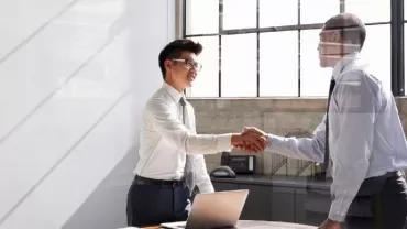 Two men in an office shaking hands by a desk with a laptop.