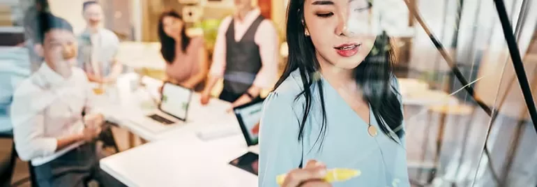 Woman writing on glass board in office meeting with colleagues watching.