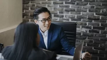 Two people in a meeting, one smiling at a laptop, in a modern office with a brick wall.