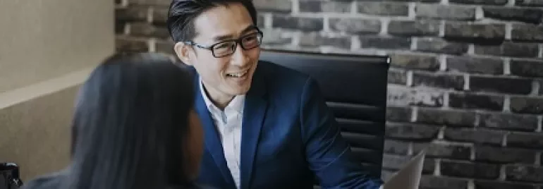 Two people in a meeting, one smiling at a laptop, in a modern office with a brick wall.