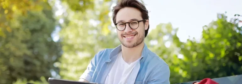 Man with glasses smiling while sitting outside with greenery in the background.