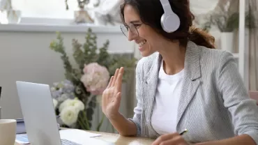 Woman with headphones using laptop, smiling and waving.