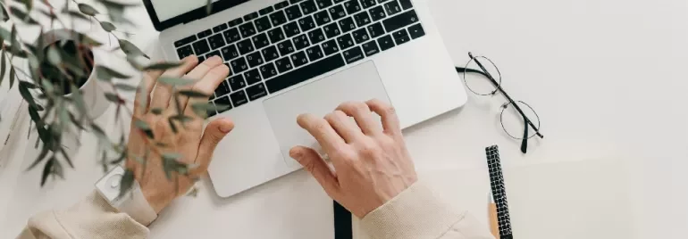 Person using a laptop on a desk with a notebook, pencil, glasses, and a plant nearby.