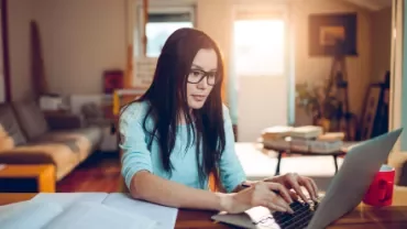 Woman with glasses working on a laptop in a cozy living room.