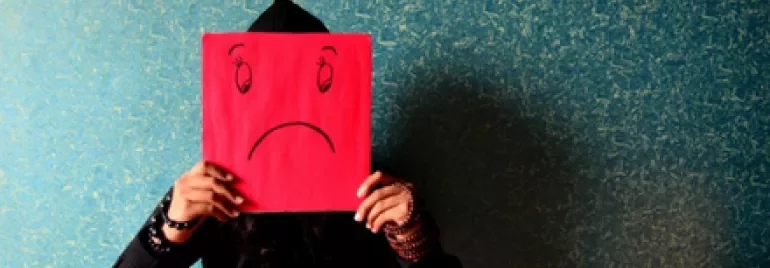 Person sitting against a textured wall holding a red paper with a sad face drawn on it.