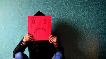 Person sitting against a textured wall holding a red paper with a sad face drawn on it.